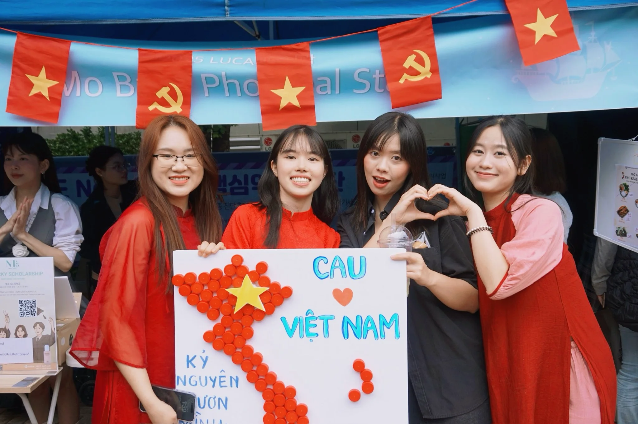 대학 축제의 베트남 유학생 부스. 베트남 국기 아래에서 'CAU VIỆT NAM' 보드를 들고 하트를 만드는 학생들.